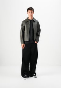 Young man standing against white background wearing dark green leather jacket, black wide-leg pants, black shirt, and black sneakers with white soles.