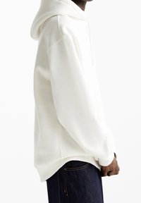 Persoon in een effen witte capuchontrui en donkerblauwe spijkerbroek, van de zijkant weergegeven met de handen voor zich samengevouwen.