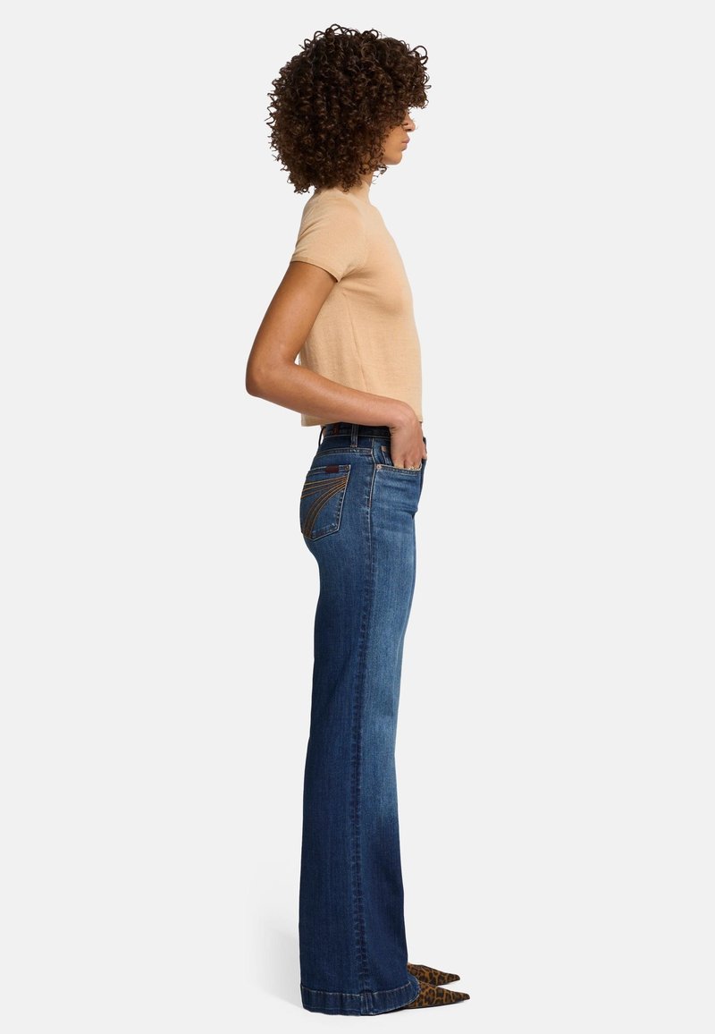 Light brown fitted top paired with blue flared jeans featuring contrasting stitching and a pocket design. Textured fabric and pointed shoes visible.