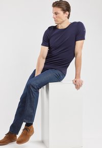 T-shirt a maniche corte di colore navy in tessuto liscio, abbinata a jeans blu scuro e scarpe in suede marrone. Modello seduto su un piedistallo bianco.