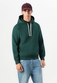 Man wearing dark green hoodie with white drawstrings, light blue jeans, and a dark red corduroy cap, standing against a plain white background.