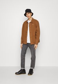 Brun knapp-up jakke, hvit T-skjorte, grå jeans, svart bøttehatt og svarte sko med gummisåle. Merkbar Nike-logo på jakken.