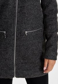 Dunkelgrauer Wollmantel mit Reißverschluss vorne, zwei seitlichen Reißverschlusstaschen und strukturiertem Stoff für Wärme. Lange Ärmel mit enganliegenden Bündchen.