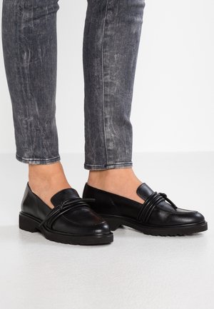 Voeten met zwarte leren loafers met decoratieve veters, gecombineerd met grijze acid-wash strakke jeans tegen een effen witte achtergrond.