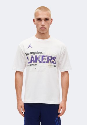 Fiatal férfi rövid szőke hajjal, fehér Los Angeles Lakers pólóban és tengerészkék sport rövidnadrágban, egyszínű háttér előtt áll.