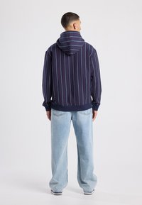 Navy raidoituksellinen huppari, jossa on nauhakuorinen huppu, yhdistettynä vaaleansinisiin väljät farkkuihin. Sisältää ribattuja hihansuita ja helman, rento istuvuus.