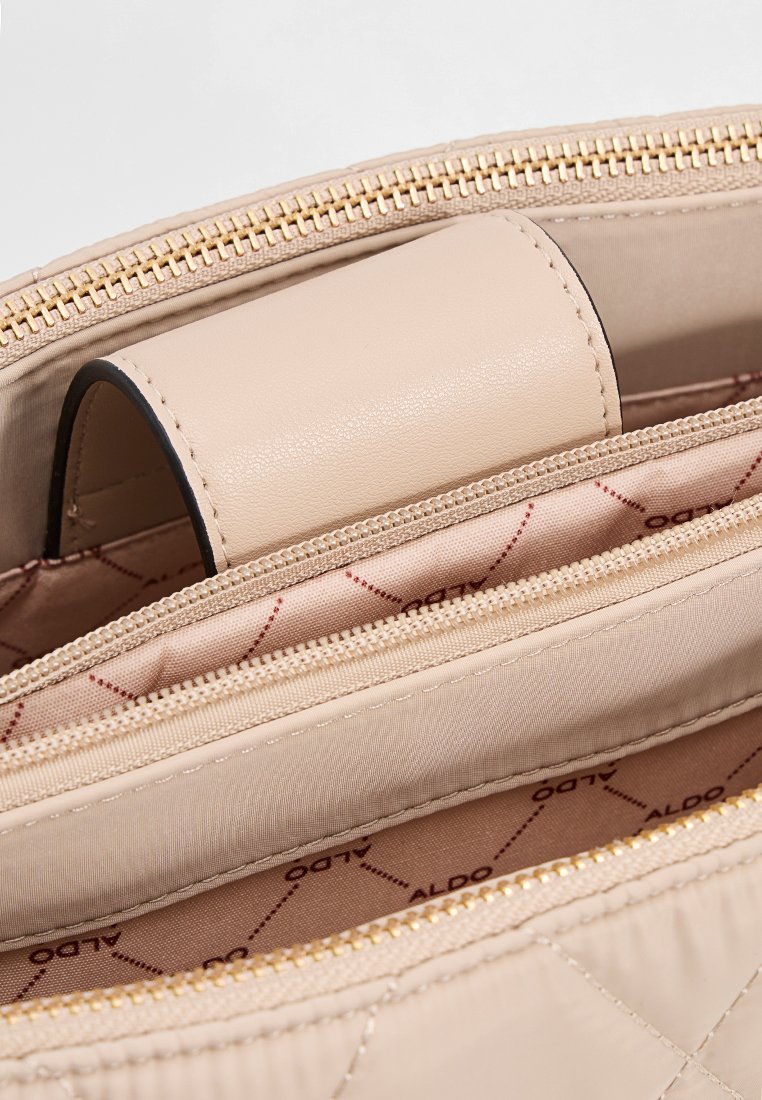 Intérieur d'un sac à main matelassé beige avec une fermeture éclair dorée, des accents en cuir lisse et une doublure en tissu imprimé de logo avec des compartiments.
