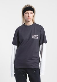 Mörkgrå bomull t-shirt med tryckt grafik på vänster bröst, lager över en vit långärmad skjorta. Avslappnad passform, enkel design.