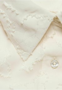 Biała bluzka z teksturowanym wzorem w kwiaty i postrzępionymi brzegami. Posiada klasyczny kołnierzyk oraz okrągły, przezroczysty guzik. Wykonana z miękkiego materiału o subtelnym połysku.