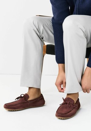 Person sidder og justerer lysegrå bukser, iført bordeaux ruskindsloafers og en mørkeblå sweater, mod en hvid baggrund.