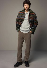 Brede gebreide trui met een kleurrijk patroon, grijze geribde shirt, bruine corduroy pantalon en zwarte veterschoenen. Neutrale achtergrond, staande pose.