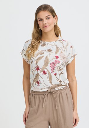 Kvinde iført hvid kortærmet top med beige og røde blomster, kombineret med lysebrune bukser med snøre, stående mod hvid baggrund.