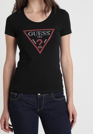 Kvinde iført en tætsiddende sort T-shirt med et rødt trekantet Guess-logo og mørkeblå jeans, stående mod en ensfarvet hvid baggrund.