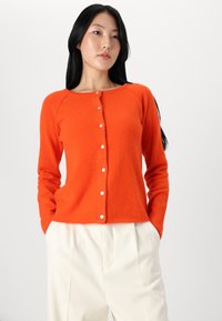 Orange cardigan made from soft material, featuring a round neckline, long sleeves, and button front with white buttons. Worn over cream trousers.
