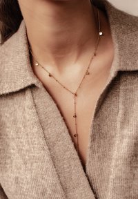 Collier doré à plusieurs couches avec des pendentifs circulaires, présentant des maillons de chaîne délicats et un design texturé, porté sur un pull en tricot brun clair.