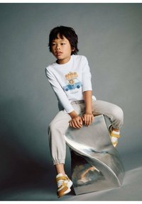 Lapsi valkoisessa pitkähihaisessa paidassa, jossa on karhu- ja auton graafinen kuva, istuu modernilla metallituolilla harmaata taustaa vasten.