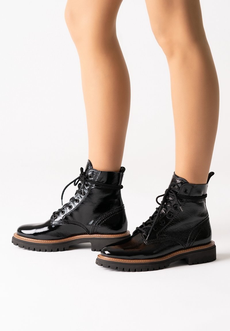 Bottes basses noires brillantes à lacets avec semelles épaisses bordées de brun, portées sur des jambes nues, sur fond blanc.