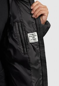 Schwarze Puffjacke mit gerippter Textur im Innenbereich, ausgestattet mit einem genähten Etikett, auf dem steht: "Eine warme Umarmung aus Stoff."