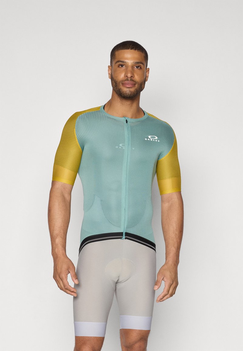 Kortermet sykkeltrøye i lyseblått med gule ermer, med glidelås foran og strukturert stoff. Kombinert med grå shorts, designet for ytelse.