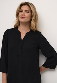 Chemise noire à manches longues avec un col et trois boutons. Tissu léger, texture lisse et coupe légèrement ample.