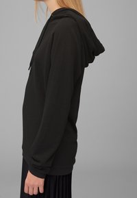 Czarna bluza z kapturem z miękkiej, gładkiej tkaniny; luźny krój z długimi rękawami i ściągaczami; noszona na plisowanej czarnej spódnicy.