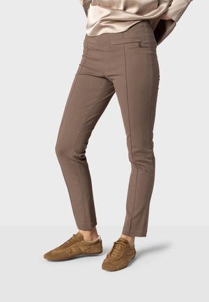 Person trägt eng anliegende taupefarbene Hose mit vorderen Nahtdetails und braune Wildledersneaker, steht vor hellem Hintergrund.