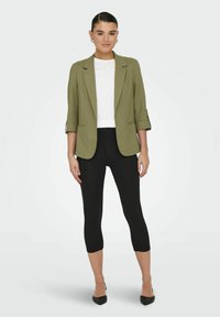 Blazer verde oliva con maniche arrotolate, top bianco aderente, leggings neri a culotte e décolleté neri a punta. Design semplice e vestibilità su misura.