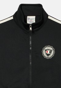 Svart treningjakke med høy krage, frontlås og hvite side-striper. Har en sirkulær logo på venstre bryst med et vintage design.