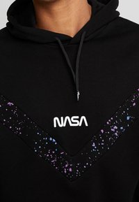 Černá mikina s kapucí s V-škovým designem a akcenty v purpurové a modré barvě. Logo "NASA" v bílé barvě je výrazně zobrazeno na hrudi.