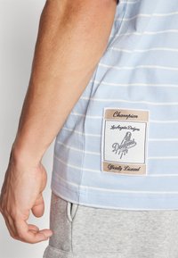 Stribet lyseblå t-shirt med et vævet mærke på siden, der indeholder teksten "Champion," "Los Angeles Dodgers," og "Officielt Licenseret."