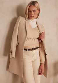 Cappotto di lana beige sopra un maglione beige chiaro, stratificato con un dolcevita bianco. Pantaloni color crema, cintura marrone e bracciali di legno.