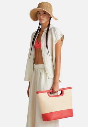 Borsa da spiaggia in paglia con rifiniture sintetiche rosse, di forma rettangolare, con manici robusti e una superficie liscia, tenuta da una persona che indossa abiti leggeri e casual.