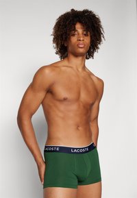 Zelene bokserice od mekane pamučne tkanine, s širokim tamnoplaviim pojasom na kojem se nalazi logo "LACOSTE". Dizajn je prilagođen i glađen.