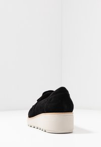 Mocassin en daim noir avec semelle plateform épaisse beige et bande de roulement crantée sur un fond blanc.