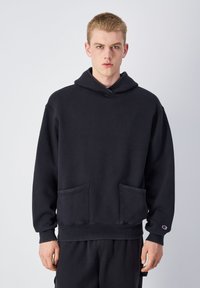 Sweatshirt noir à capuche avec une poche kangourou, des poignets et un ourlet côtelés. Fabriqué en tissu doux, il présente un patch logo sur la manche gauche.