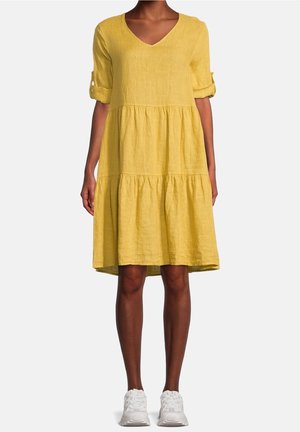 Femme portant une robe jaune ample jusqu'aux genoux avec les manches retroussées et des baskets blanches, debout devant un fond uni.