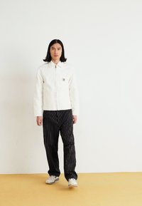 Witte ritsjack met een kraag en een vo pocket, gecombineerd met zwart gestreepte broek. Achtergrond is eenvoudig wit, vloer is geel.