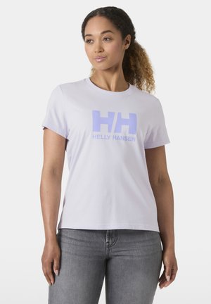 Kobieta o kręconych włosach, ubrana w białą koszulkę Helly Hansen i szare dżinsy, stoi na tle jednolitego jasnego tła.