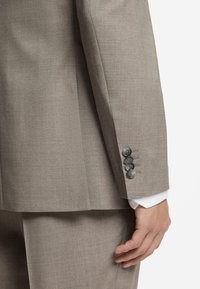 Manche de veste de costume beige avec quatre boutons gris, poignet de chemise blanche visible, et pantalon de costume beige assorti sur le bras et la main d’une personne.