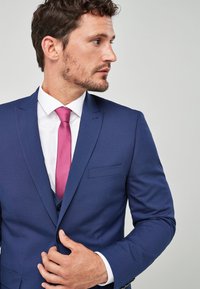 Marineblauer Anzug mit Slim Fit, ausgestattet mit einem Revers und einer einzelnen Brusttasche. Begleitet von einer rosa Krawatte über einem weißen Hemd.