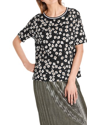 Kobieta w czarnej bluzce z krótkim rękawem, z białym i brązowym kwiatowym wzorem oraz okrągłym kołnierzykiem, zestawiona z metaliczną plisowaną spódnicą.