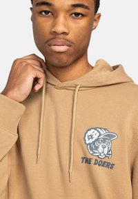 Tan hoodie made of soft fabric featuring a graphic of a dog wearing a cap, with the text "THE DOERS" below the design.