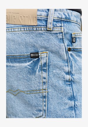 Lys blå denimjeans har en stor baglomme med et sort mærke, gul syning og en tekstureret taljeblæk med indprentet "BIG STAR."