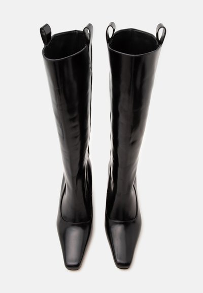 Paire de longues bottes en cuir noir avec bouts carrés et languettes en haut, présentées debout sur fond blanc.
