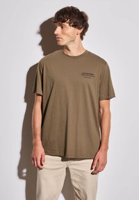 Olivově zelené bavlněné tri čko s kulatým výstřihem a krátkými rukávy, na levé části hrudi s černým textem. Model má na sobě béžové kalhoty.