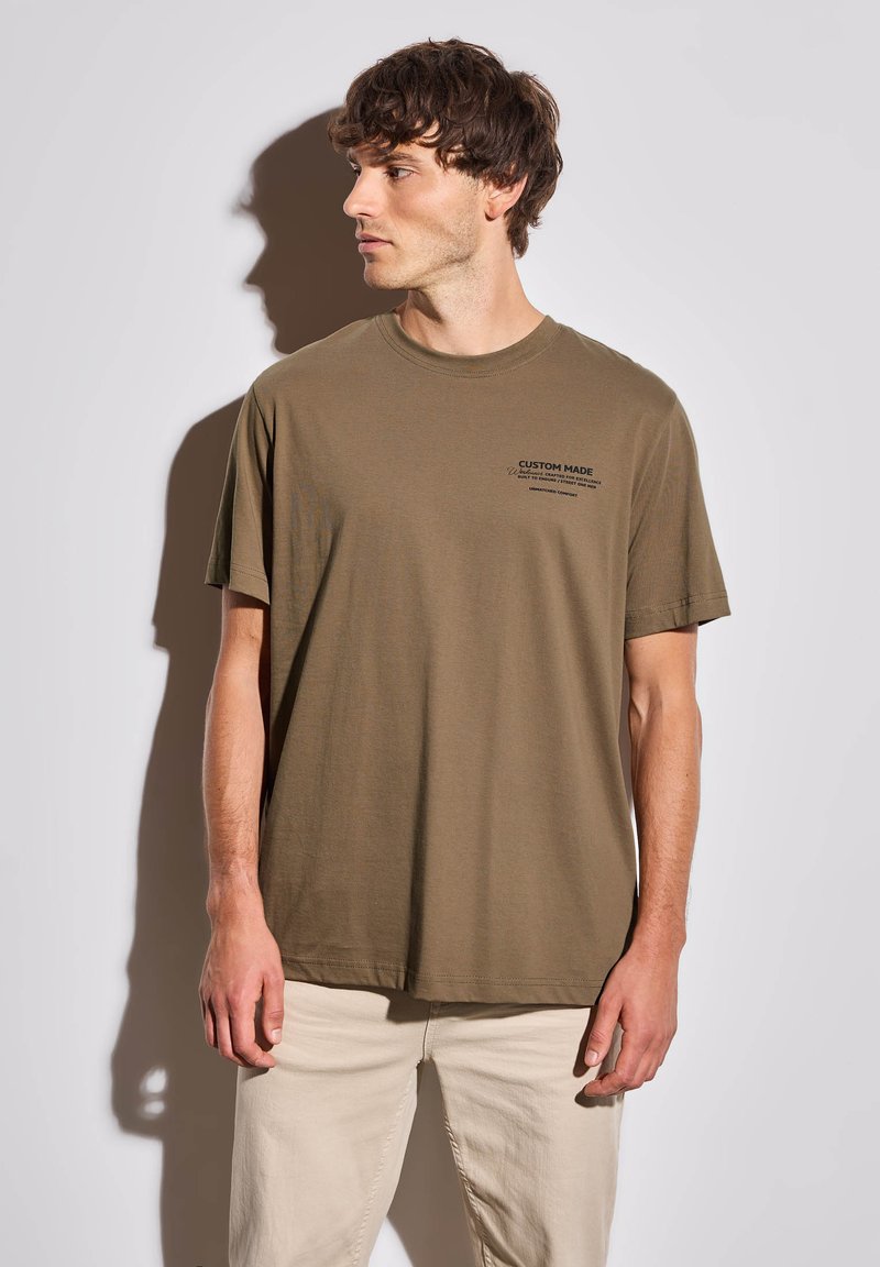 Olivově zelené bavlněné tričko s kulatým výstřihem a krátkými rukávy, na levé části hrudi s černým textem. Model má na sobě béžové kalhoty.