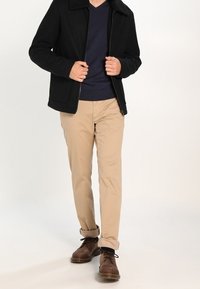 Giacca zip nera con colletto testurizzato, maglione a V blu navy, pantaloni beige e scarpe stringate marroni con occhielli metallici.