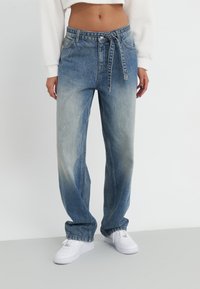 Blue denim jeans with a high waist, loose fit, light fading, and a tied belt. Paired with white sneakers on a light background.