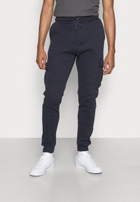 Marine cargobroek met elastische tailleband, trekkoord, zijzakken en taps toelopende benen, gecombineerd met witte sneakers op een lichte achtergrond.