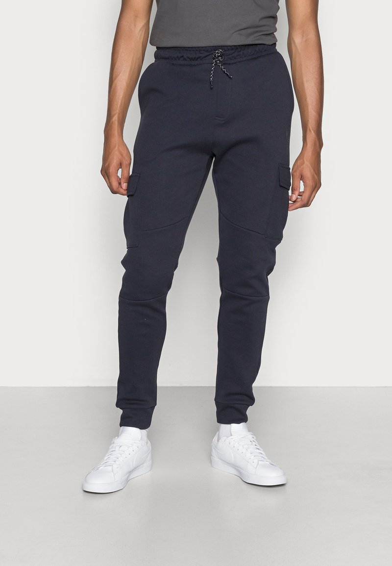 Marine cargobroek met elastische tailleband, trekkoord, zijzakken en taps toelopende benen, gecombineerd met witte sneakers op een lichte achtergrond.